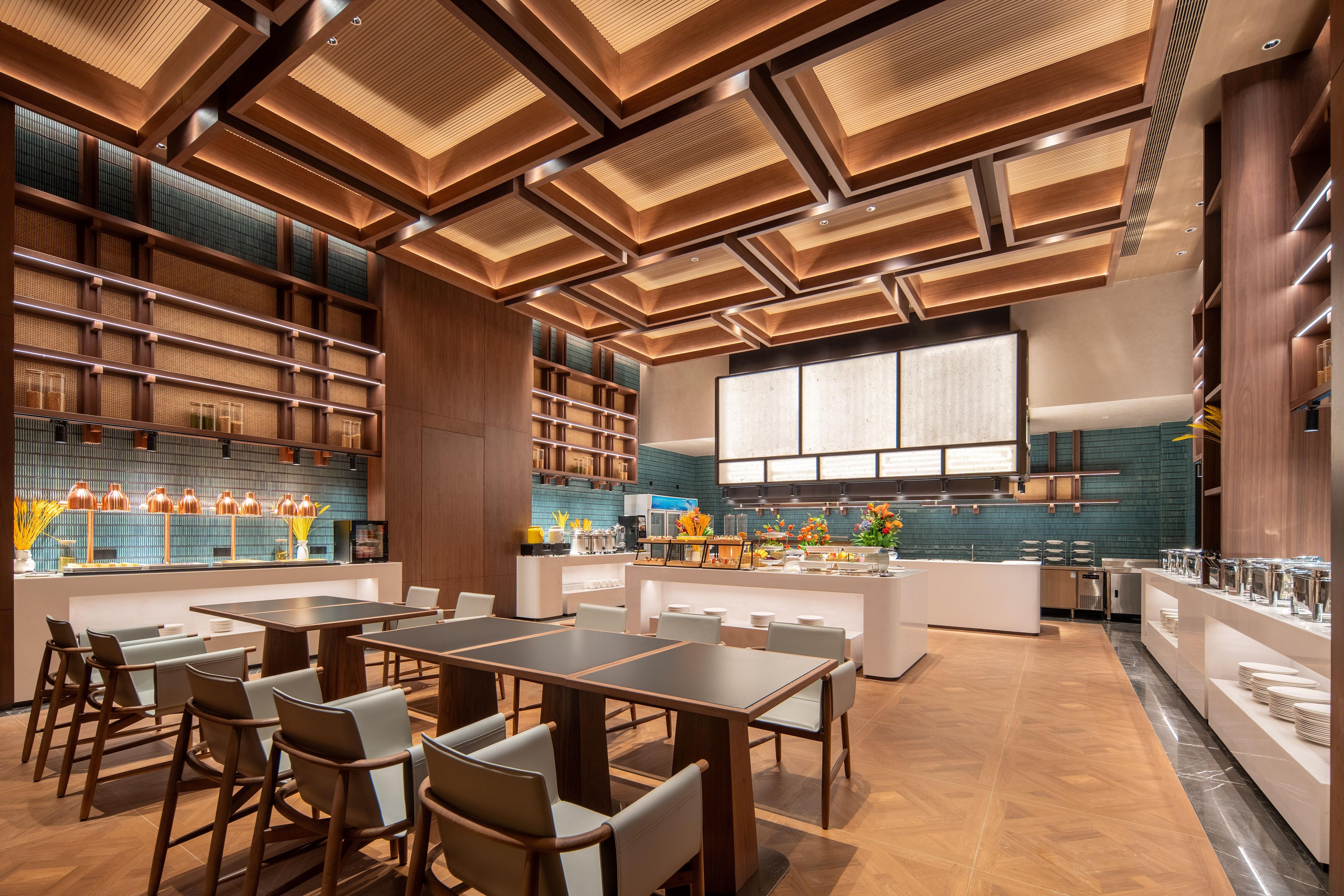 A modern dining area with wooden tables and chairs, a kitchen in the background, and a large window providing natural light.