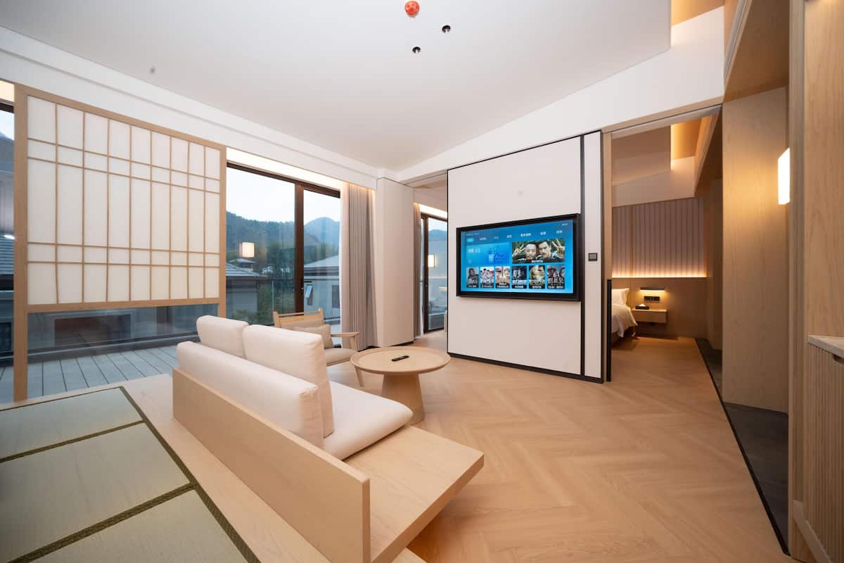 A modern hotel room with a sofa, a round table, a TV on the wall, and a view of the outdoors.