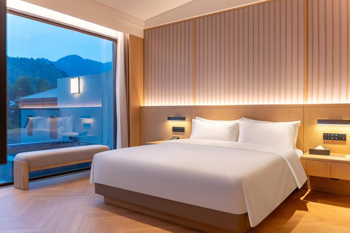 A modern hotel room with a large bed, bedside tables with lamps, a bench, and a large window offering a view of the mountains.