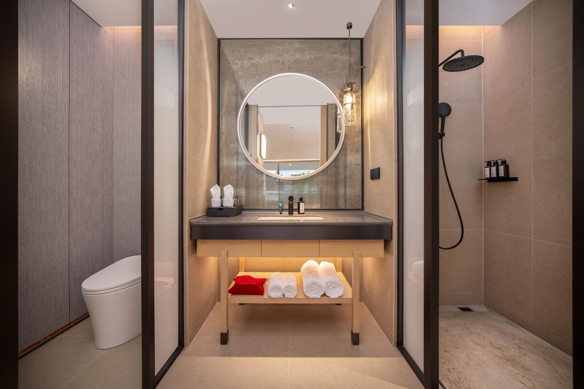 A modern bathroom with a round mirror, a sink, a shower, and neatly folded towels on a shelf.