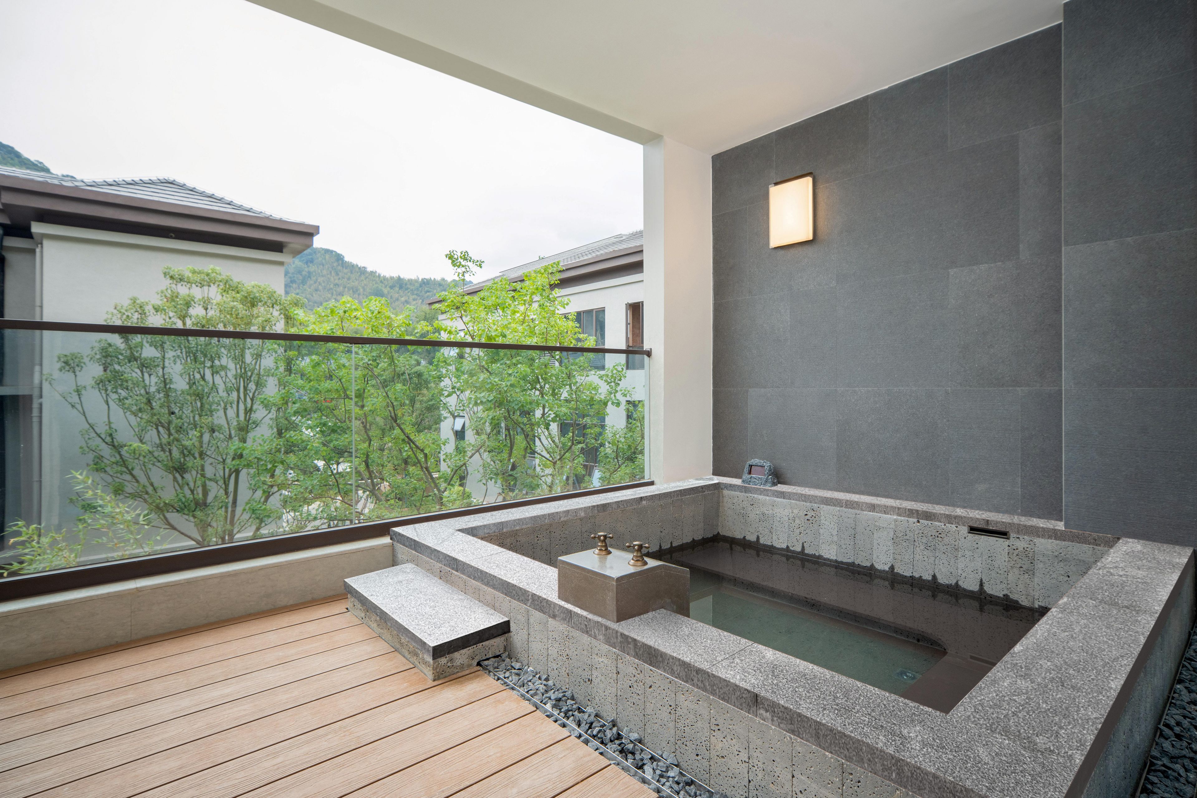 A modern spa area with a stone hot tub, wooden deck, and a view of surrounding buildings and trees.