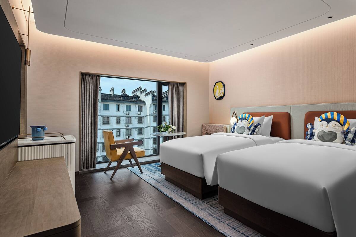 Ett hotellrum med två sängar, ett skrivbord, en stol, ett fönster med utsikt över byggnader och ett litet bord med en växt.