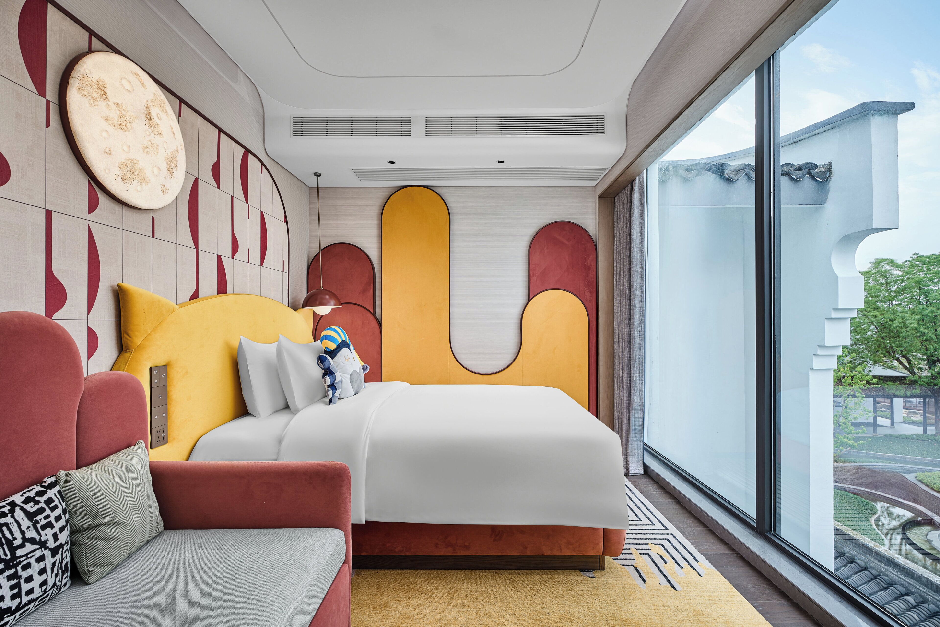 A modern hotel room with a large bed, a red sofa, and a large window offering a view of the outside.