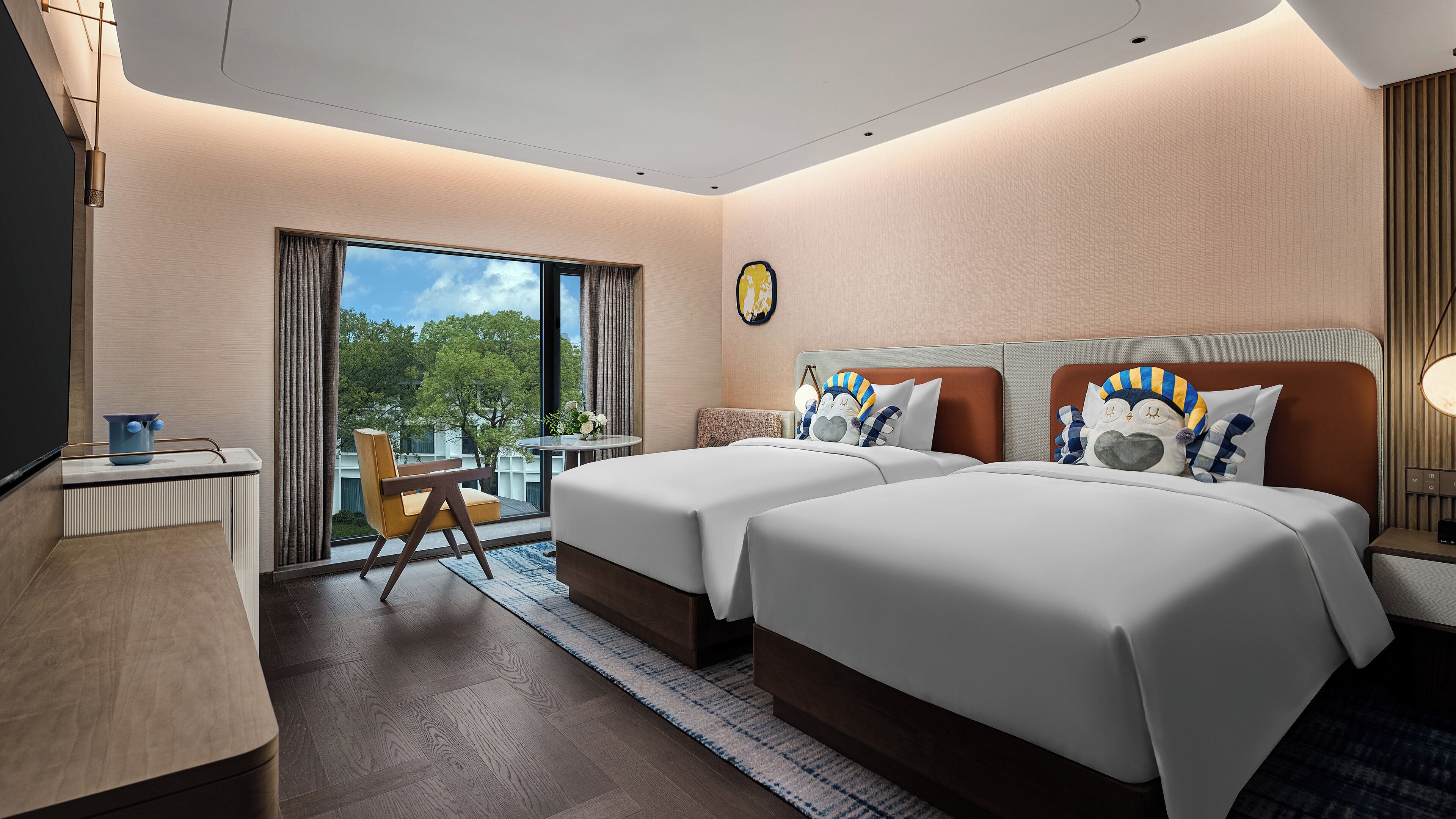 A hotel room with two beds, a desk, a chair, and a view of greenery.