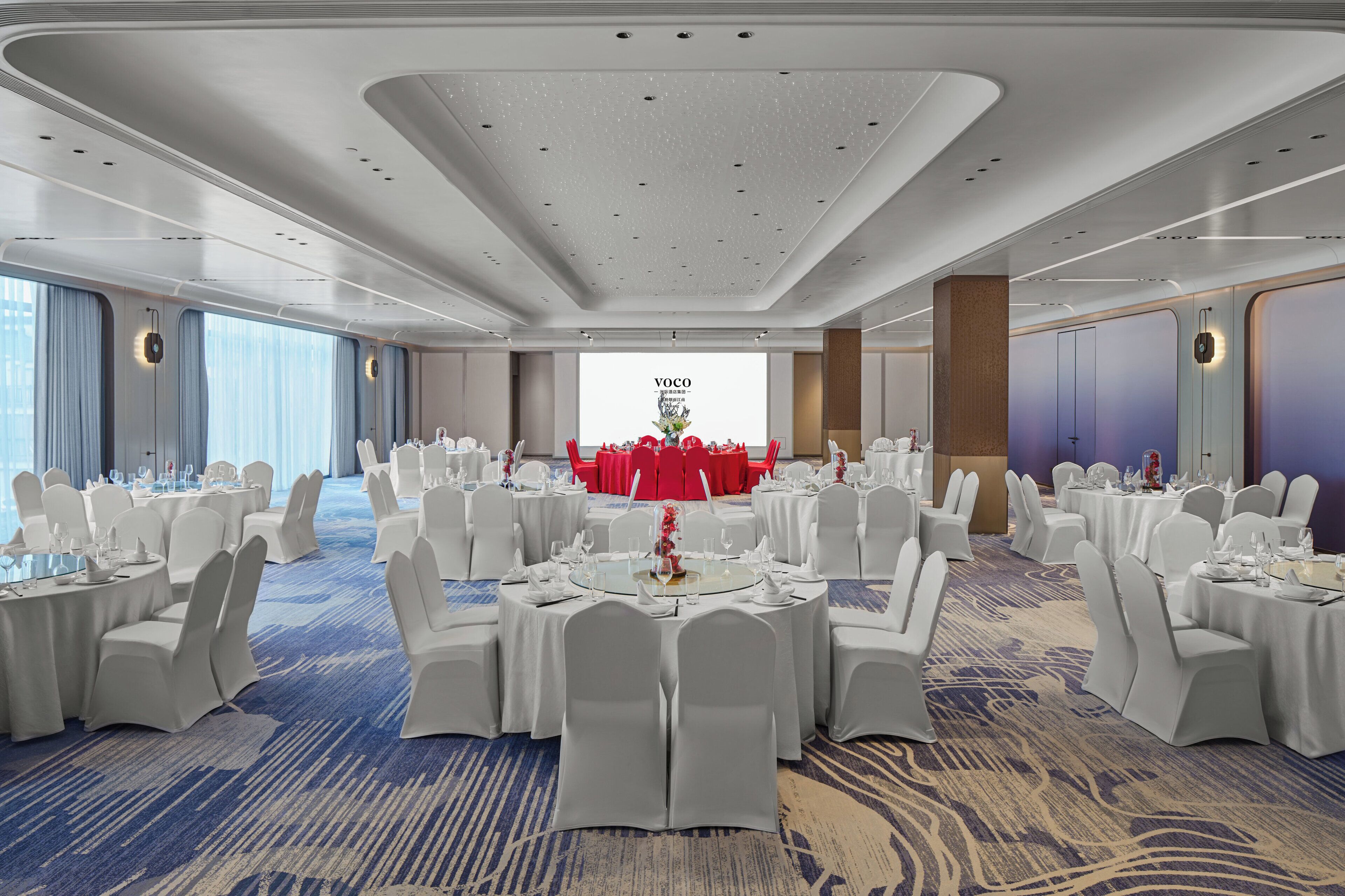 Une salle de banquet avec des tables rondes, des chaises blanches et un grand écran affichant du texte.