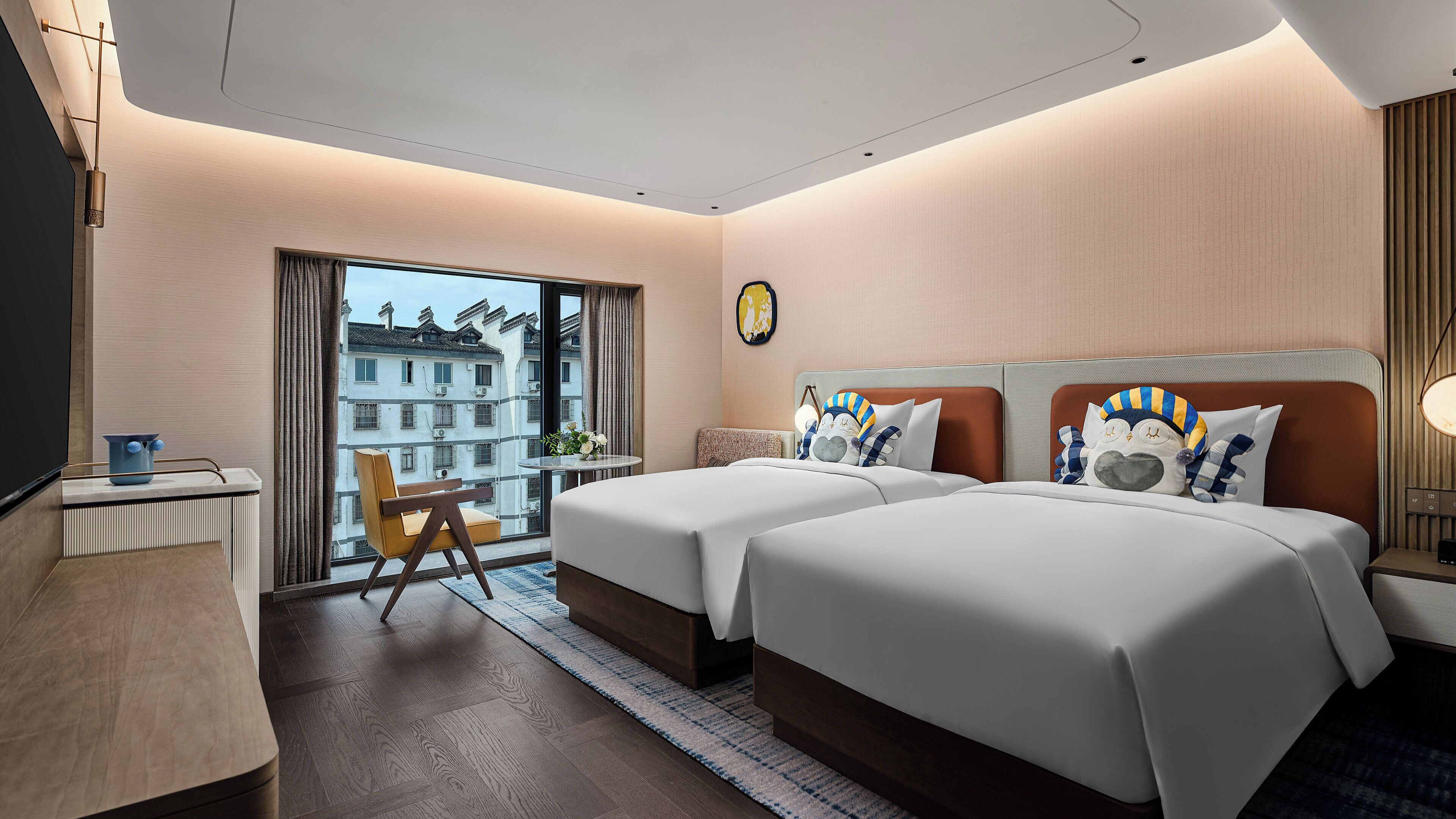 A hotel room with two beds, a desk, a chair, a window with a view of buildings, and a small table with a plant.