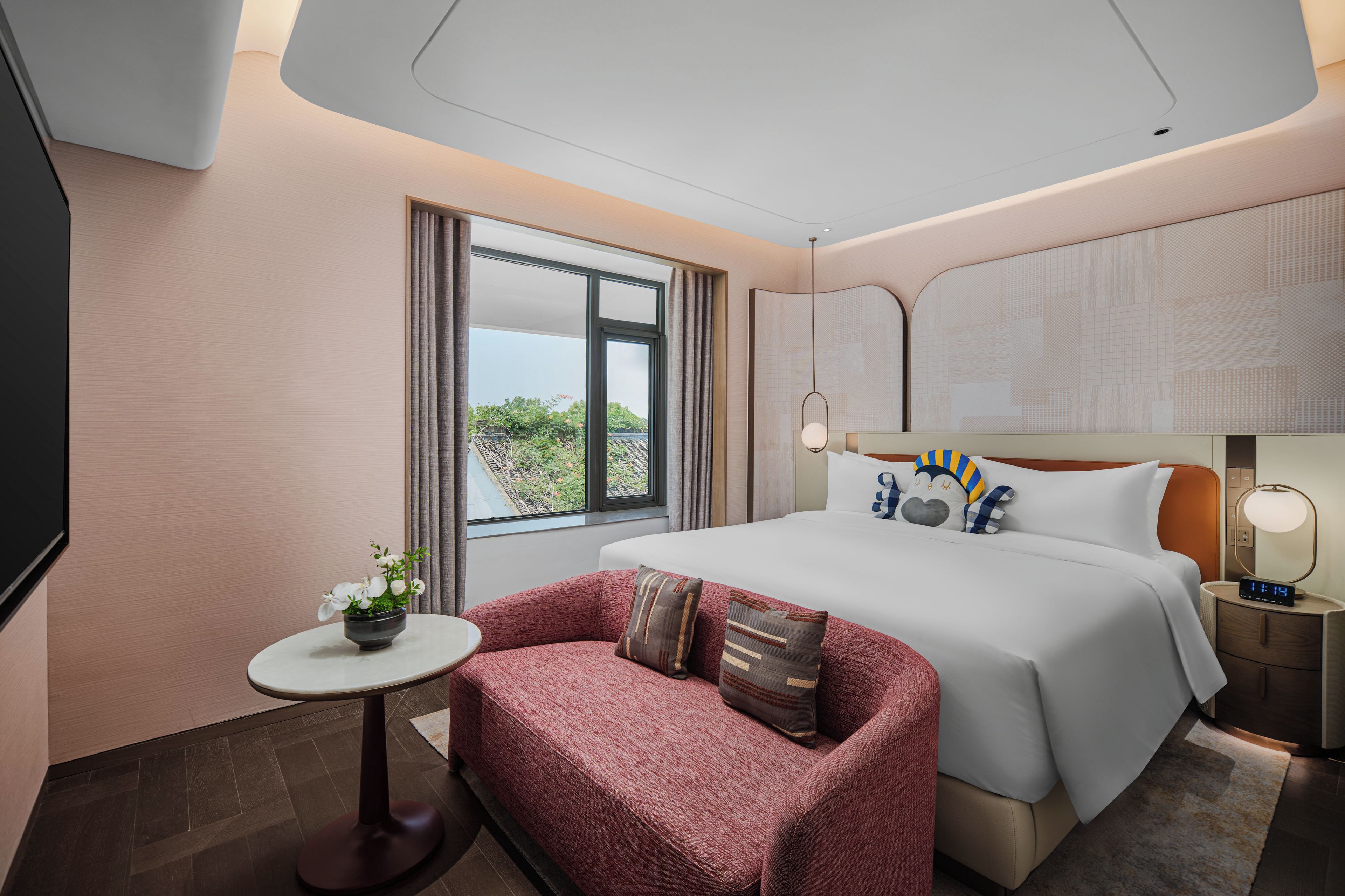 A hotel room with a bed, a sofa, a small table with a plant, a nightstand with a lamp, and a window with curtains.