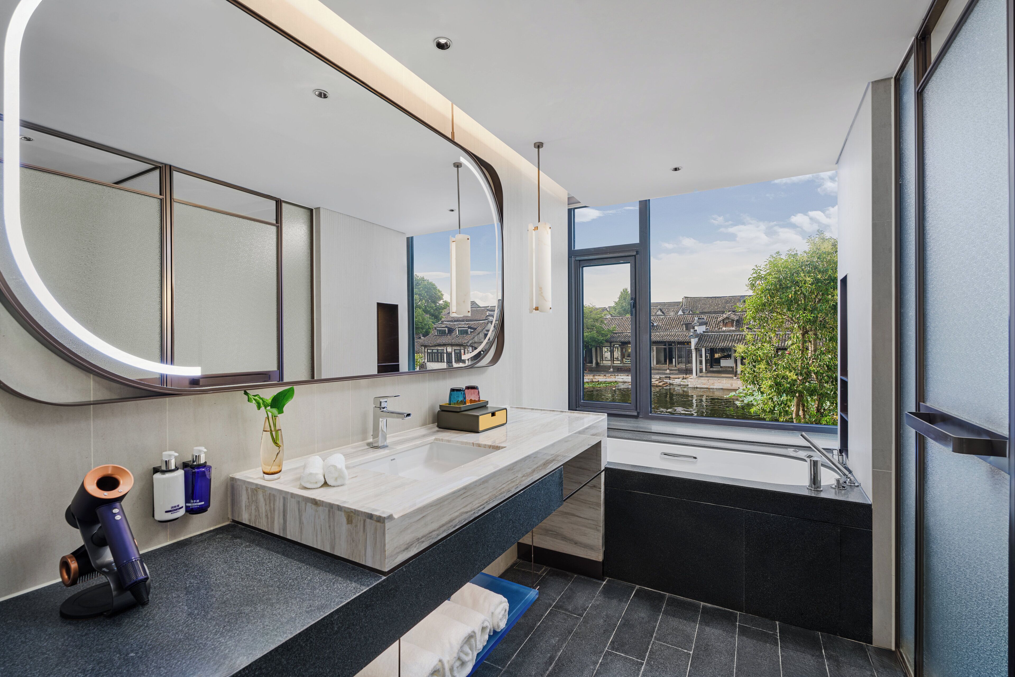 Un bagno moderno con un grande specchio, un lavandino e una finestra che si affaccia su edifici e alberi.