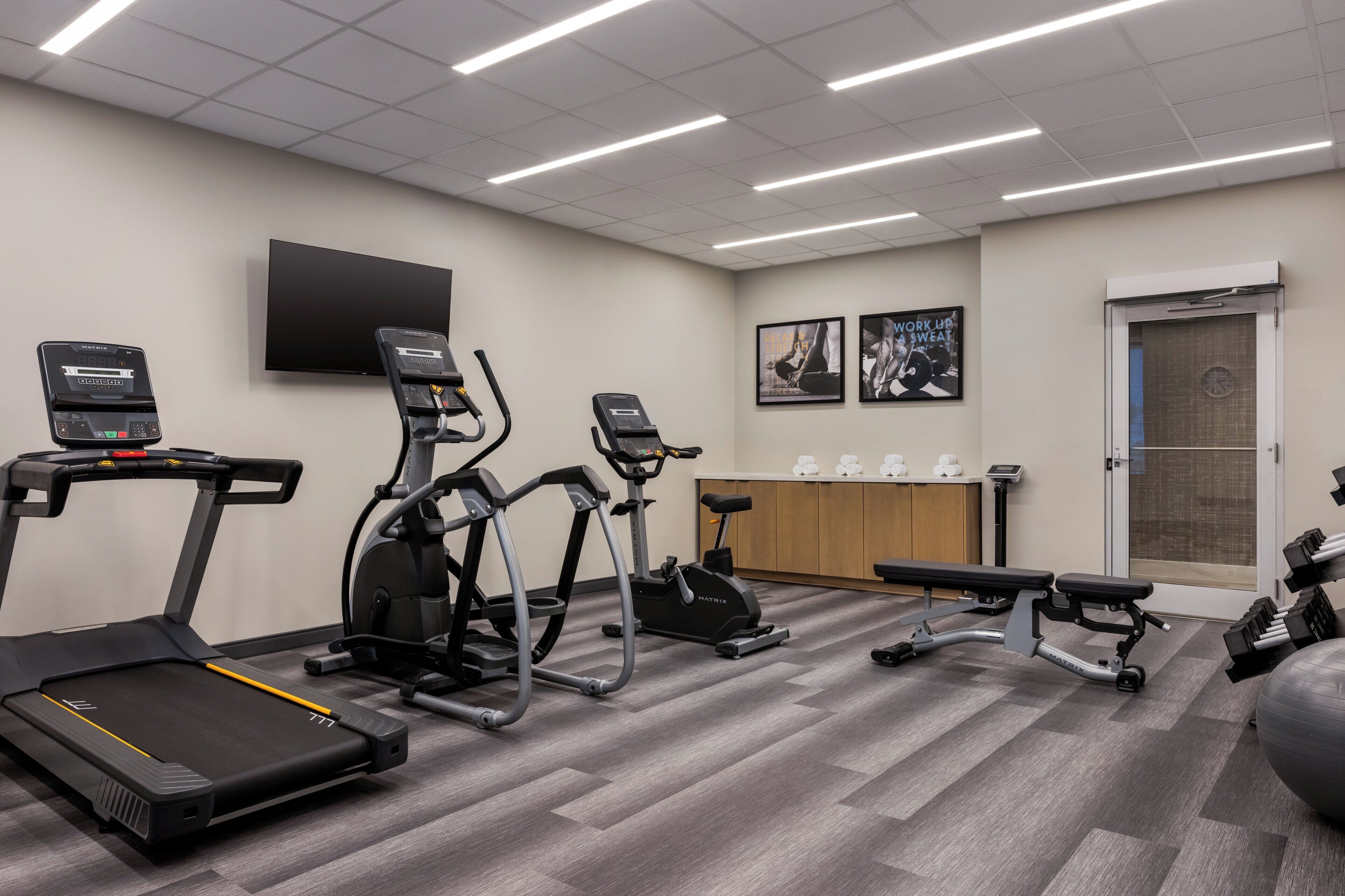 A gym with treadmills, elliptical machines, a bench, and a wall-mounted TV.