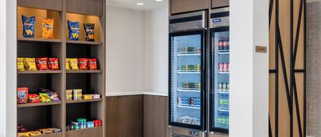 A hotel pantry with shelves stocked with snacks and a refrigerator with beverages.