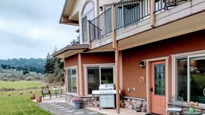 Une maison à deux étages avec un balcon, une terrasse dotée d’un barbecue et une vue sur un paysage verdoyant.