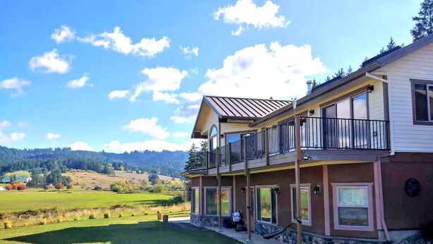 Une maison à deux étages avec un balcon, une pelouse et une vue sur une zone boisée.
