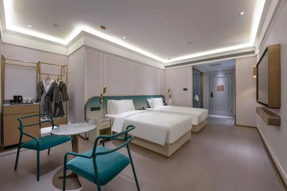 Habitación de hotel con cama, dos mesitas de noche, una silla, una mesita pequeña, un espejo y televisor.