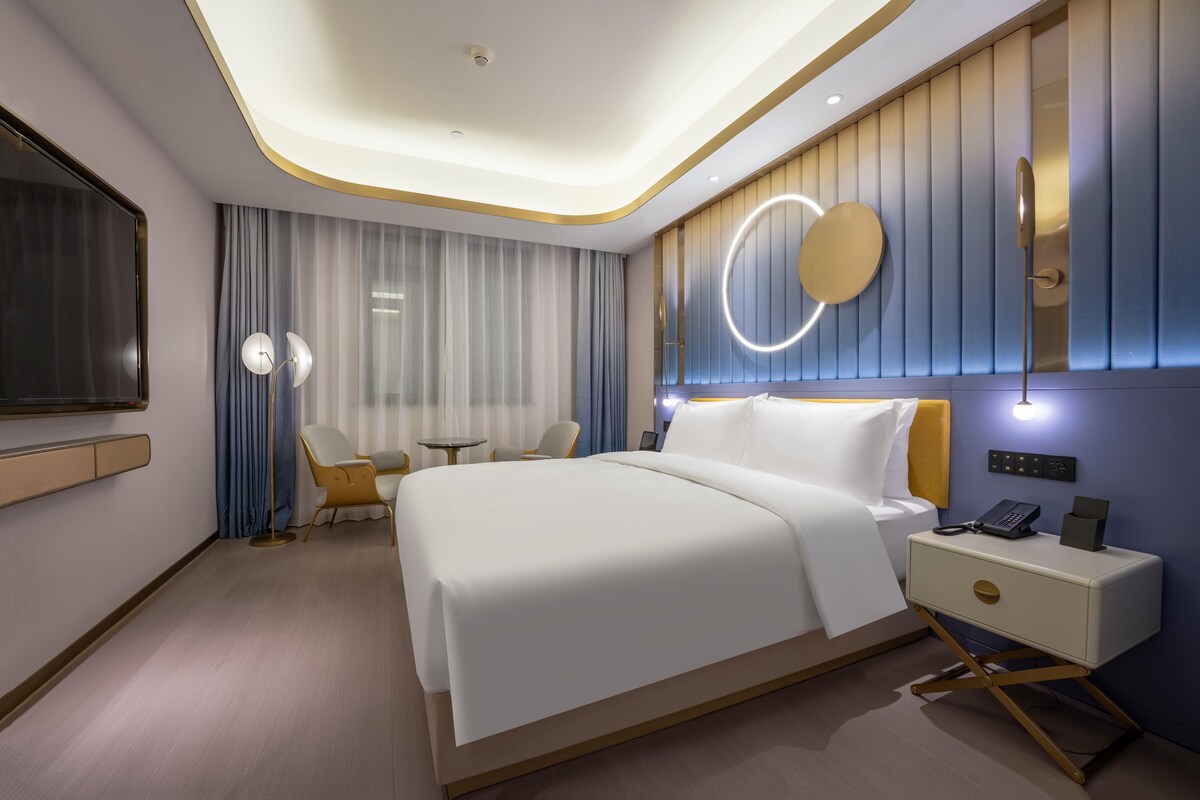 Quarto de hotel moderno com uma cama grande, mesa de cabeceira e um espelho redondo na parede.