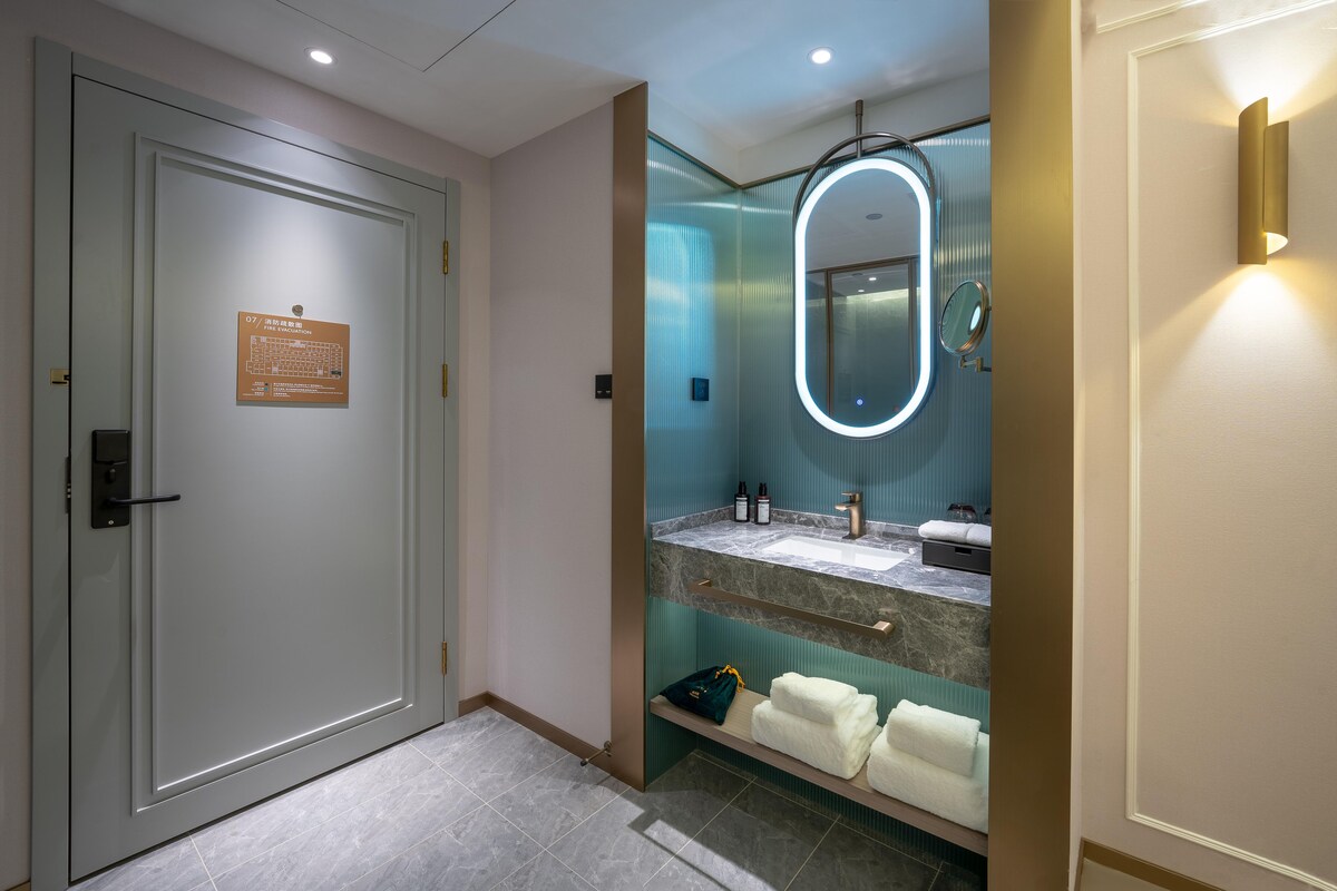 Un baño de hotel con una puerta blanca, un espejo con luz, un lavabo y un perchero para toallas.
