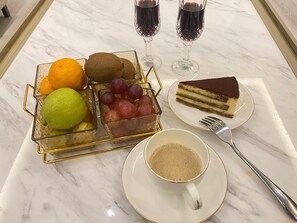 Una mesa de mármol con una cesta de frutas, una porción de pastel, dos copas de vino tinto, una taza de café y un tenedor.