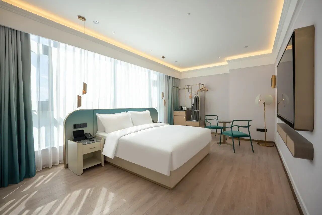 Moderni hotellihuone, jossa on suuri sänky, yöpöytä puhelimella, työpöytä tuolilla ja seinälle asennettu televisio.