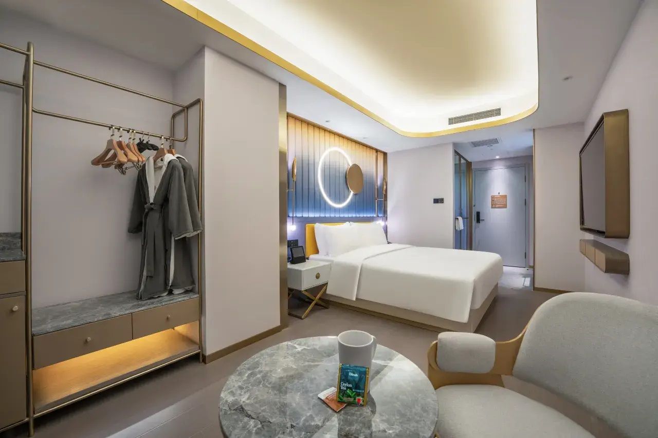 A modern hotel room with a bed, bedside table, a round coffee table, a seating area, a wardrobe with hanging clothes, and a circular light fixture on the wall.