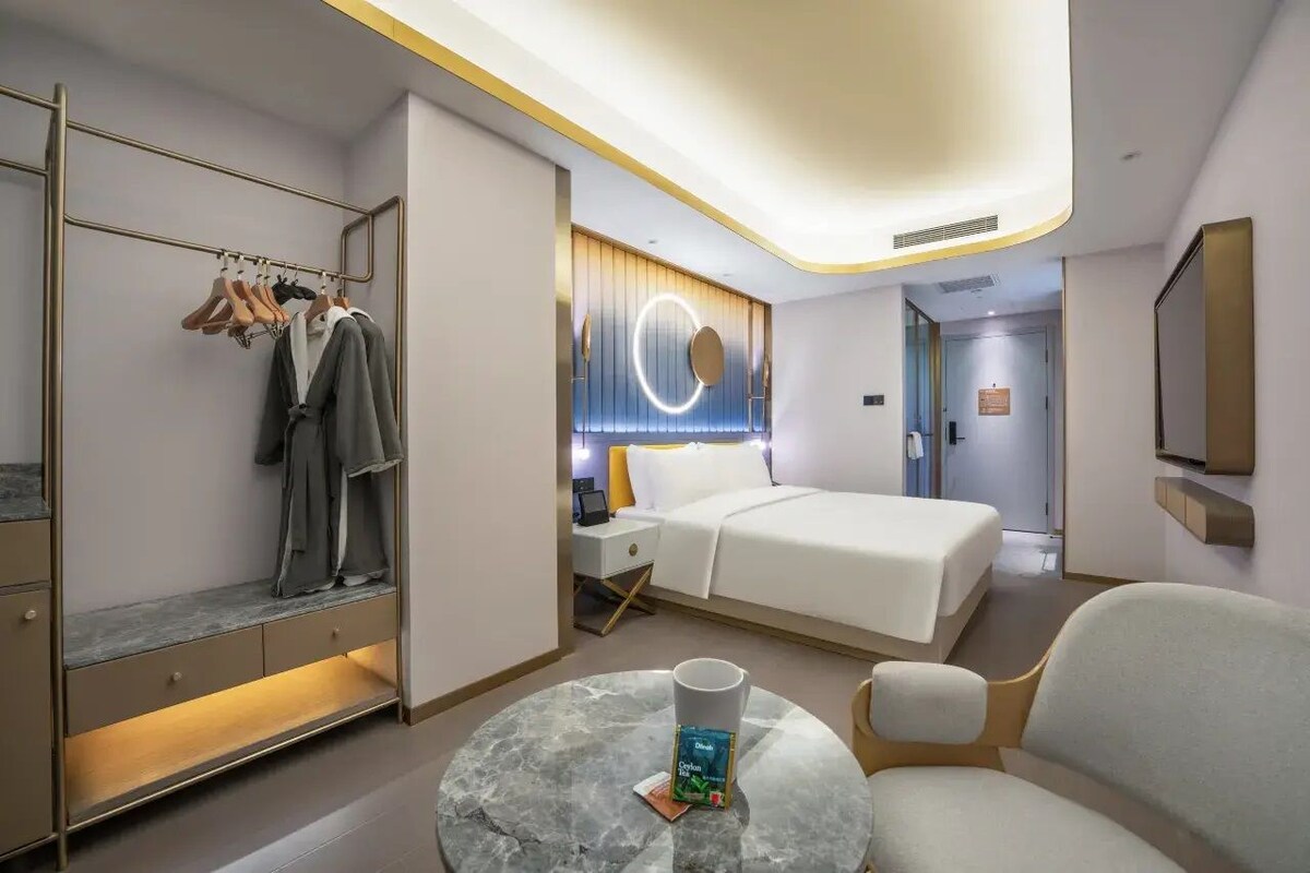 Una habitación de hotel moderna con una cama, mesita de noche, una mesa de centro redonda, un área de descanso, un armario con ropa colgada y un aplique de luz circular en la pared.