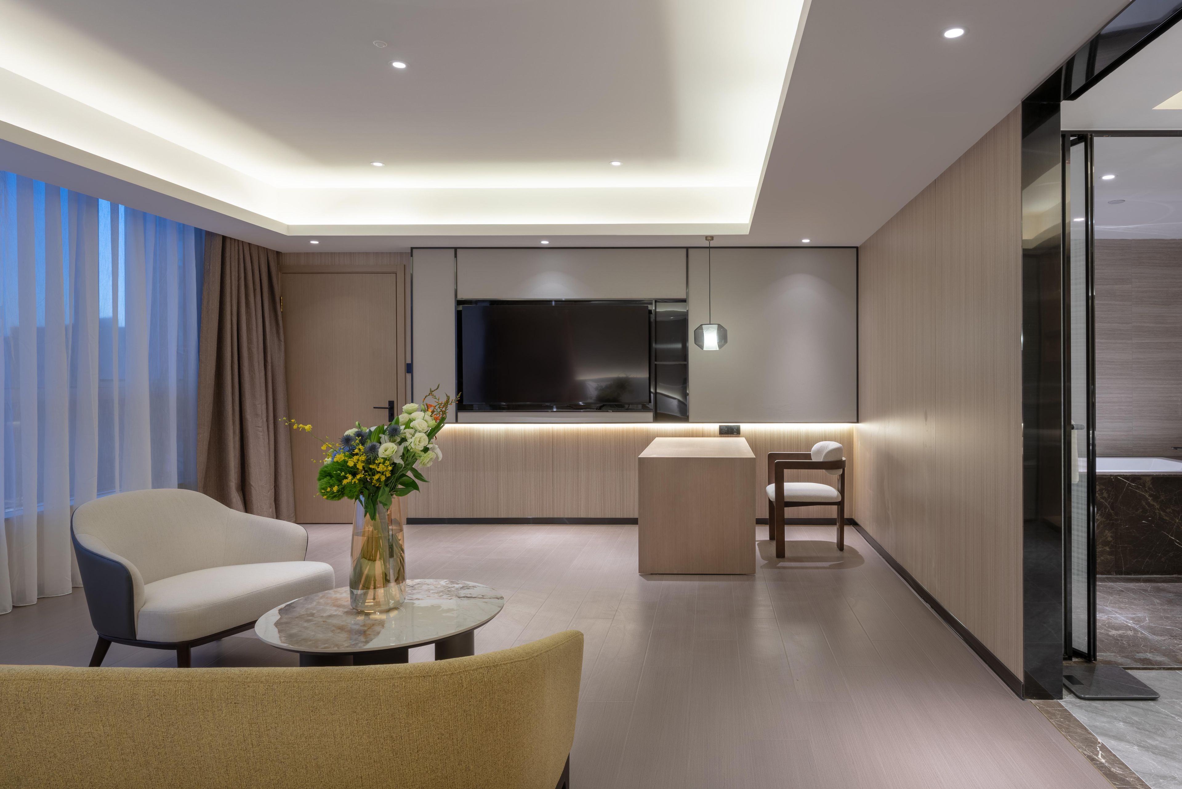 A modern hotel room with a sofa, a round coffee table, a TV, and a glass partition.