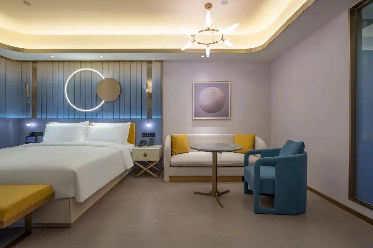 A modern hotel room with a bed, bedside table, nightstand, sofa, armchair, and a small round table.