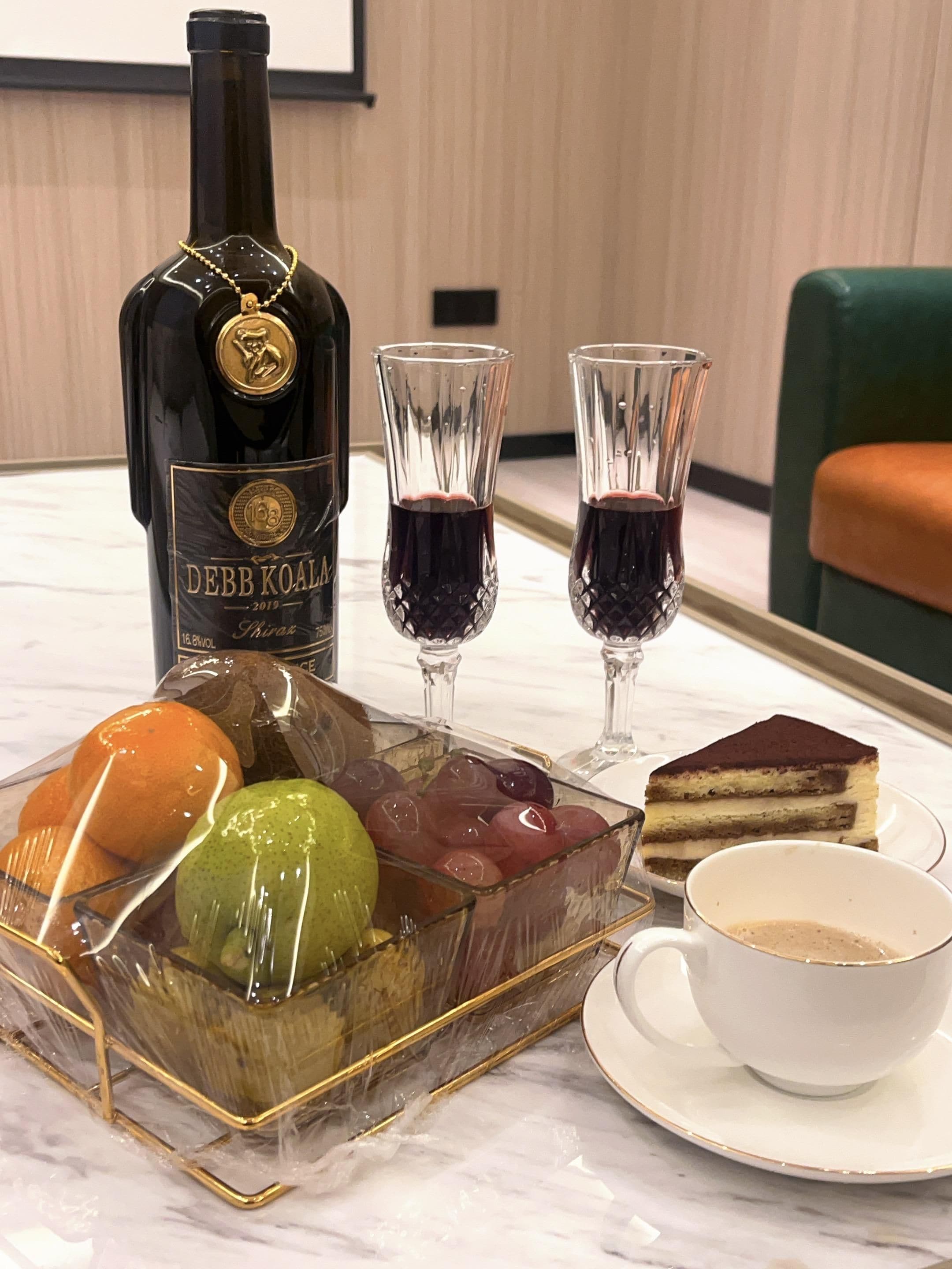 A bottle of Debb Koala wine, two wine glasses, a fruit bowl with oranges and grapes, a slice of cake, and a cup of coffee on a marble table.