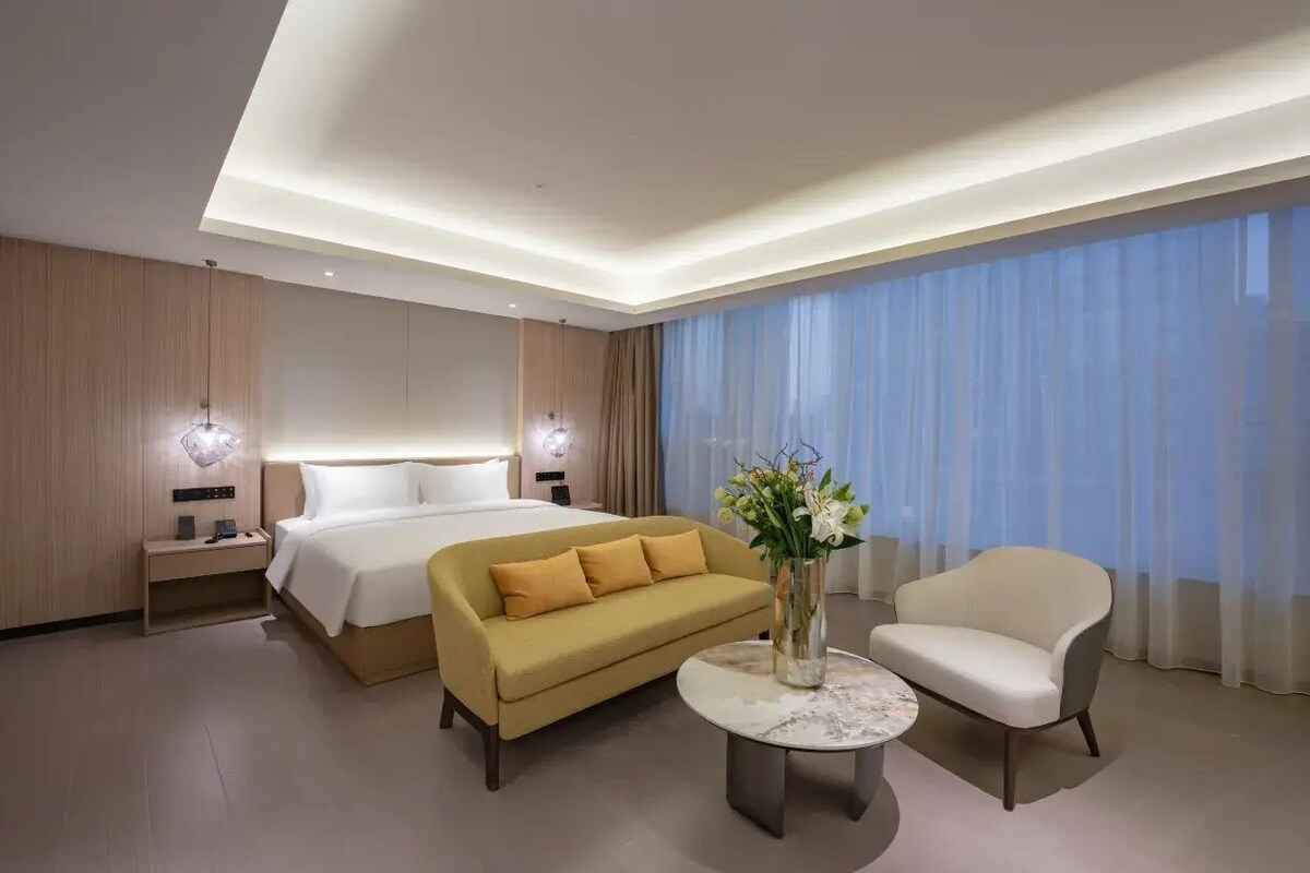 Una habitación de hotel moderna con una cama, un sofá, una silla, una mesita y un florero con flores.