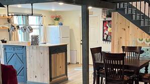 A modern kitchen with wooden cabinets, a central island, and hanging pots. There is a dining area with a table and chairs, and a staircase leading to another level.