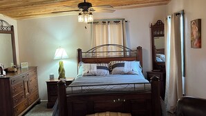 A bedroom with a wooden bed, a nightstand with a lamp, a dresser, a chest of drawers, a ceiling fan, and a window with curtains.