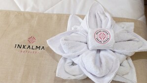 A neatly folded white towel with a logo on it, placed on a beige fabric surface with the text "INKALMA HOTELS" visible. - Inkalma sede Don Bosco (Arequipa)