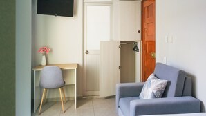 A compact room with a desk, a chair, a grey sofa, and a television mounted on the wall. - Inkalma sede Don Bosco (Arequipa)
