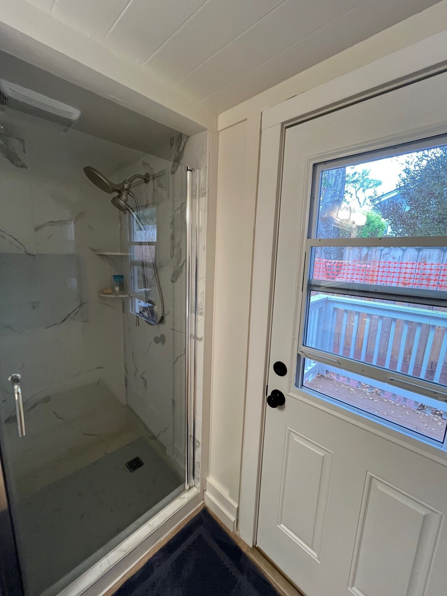 A glass-enclosed shower with a handheld showerhead, a glass door, and a view of a wooden deck outside.