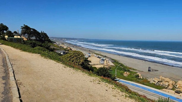 Une route côtière bordée d’une plage, avec des voitures garées et une vue dégagée sur l’océan.