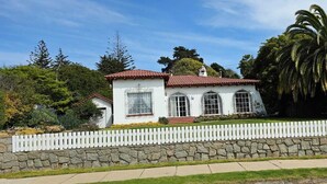 Une maison blanche avec un toit rouge, entourée d’un mur de pierre et d’une clôture en bois peinte en blanc.
