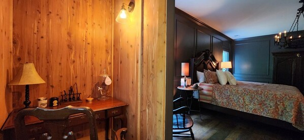 A room with a wooden wall, a desk with a lamp, a chair, a bed with a patterned bedspread, and a chandelier.