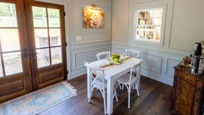 Un coin repas avec une table et des chaises blanches, un lustre, un tableau au mur et une fenêtre avec des stores.