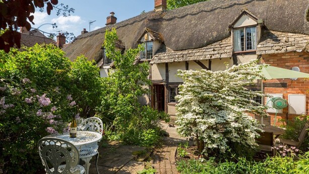 A traditional thatched-roof cottage with a garden patio, a table with chairs, and blooming flowers. - Pass the Keys | Dara Cottage | 16th Century Thatched Cottage (Mickleton)