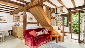 A cozy room with a red sofa, wooden beams, a staircase, and a view of greenery outside. - Pass the Keys | Dara Cottage | 16th Century Thatched Cottage (Mickleton)