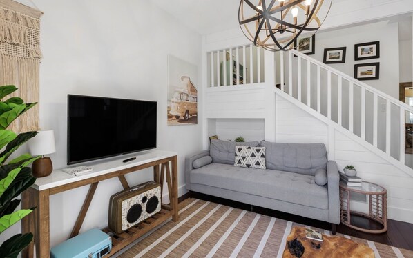 A living room with a flat-screen TV on a wooden console, a grey sofa with patterned cushions, a staircase with white railings, and a macrame wall hanging.