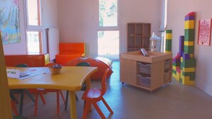 A brightly lit room with a yellow table, orange chairs, a wooden play area, and a window with a view of trees. - Beautiful Family Apartment in Amboise (Amboise)