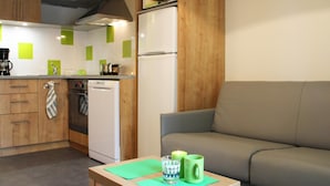 A compact kitchen with wooden cabinets, a white dishwasher, and a microwave. A small table with a green placemat holds a glass and a mug. A grey sofa is in the background. - Beautiful Family Apartment in Amboise (Amboise)