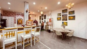 A dining area with white chairs and a round table, a bar area with a Coca-Cola refrigerator, and artwork on the walls.