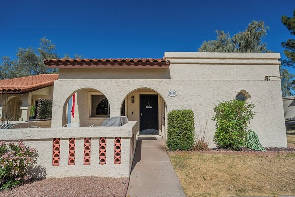 A stucco building with a tiled roof, a front door with a 'Welcome' sign, and a small garden area with shrubs.