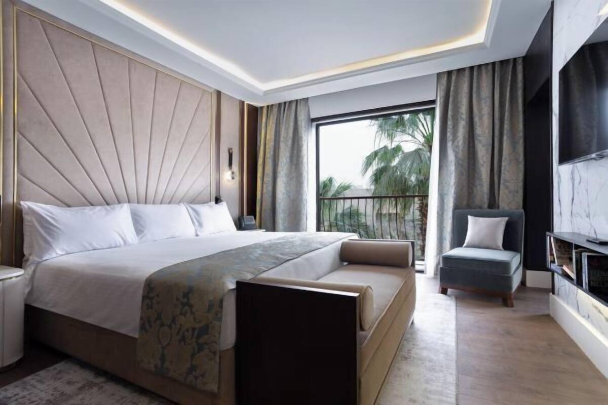 A modern bedroom with a large bed, a sofa, a TV, and a balcony with a view of palm trees.