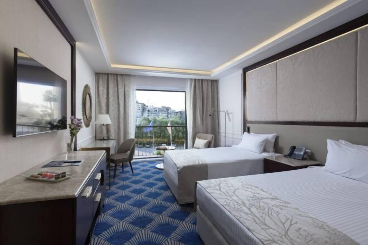 A hotel room with two beds, a desk, a chair, a TV, and a balcony with a view of buildings.