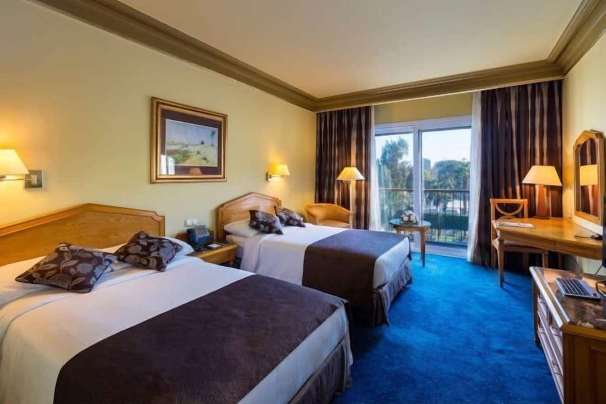 A hotel room with two beds, a desk, a chair, a lamp, a painting, and a view of the outdoors.