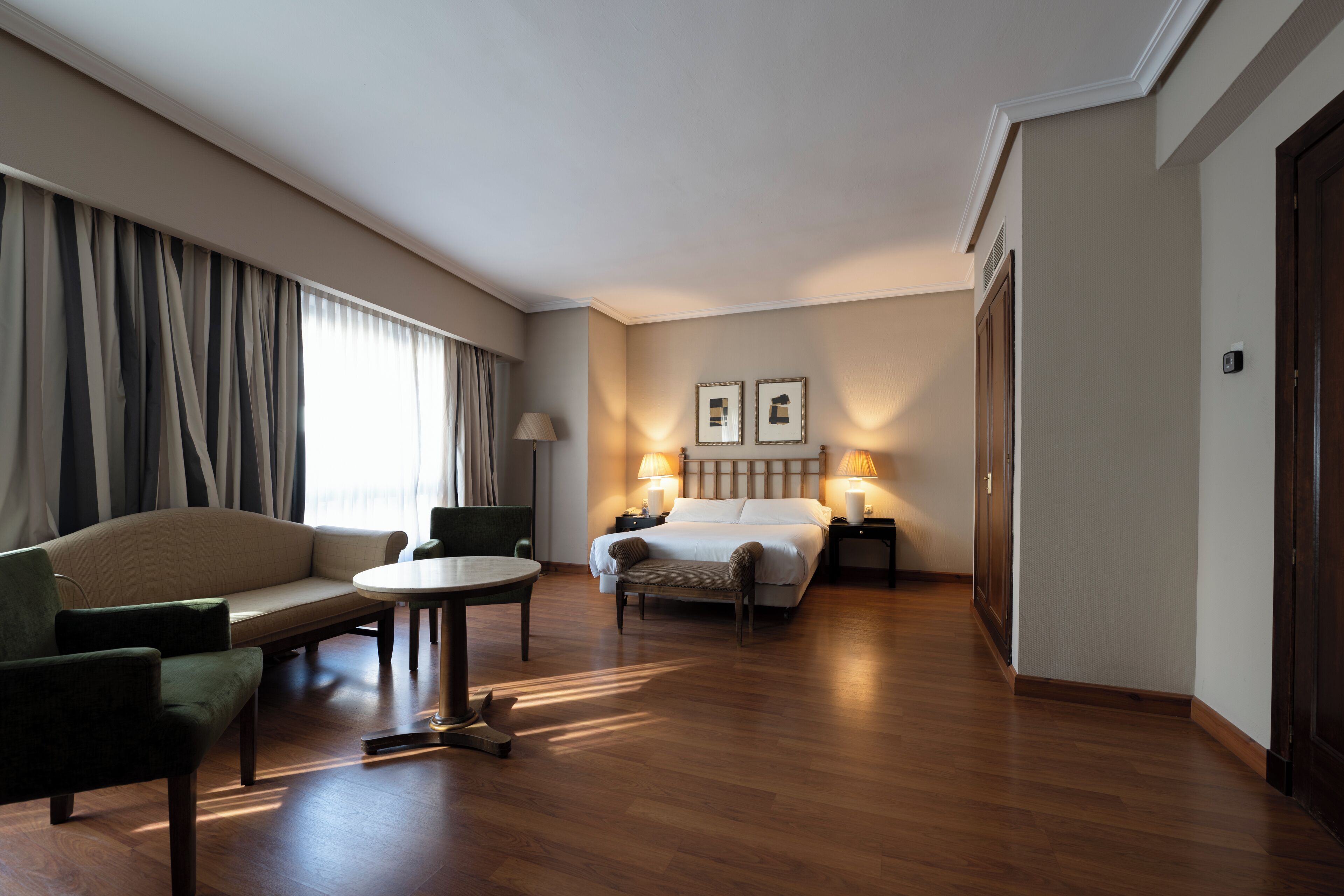 A hotel room with a bed, a sofa, a small table, and a chair. There are two lamps on the bedside tables.