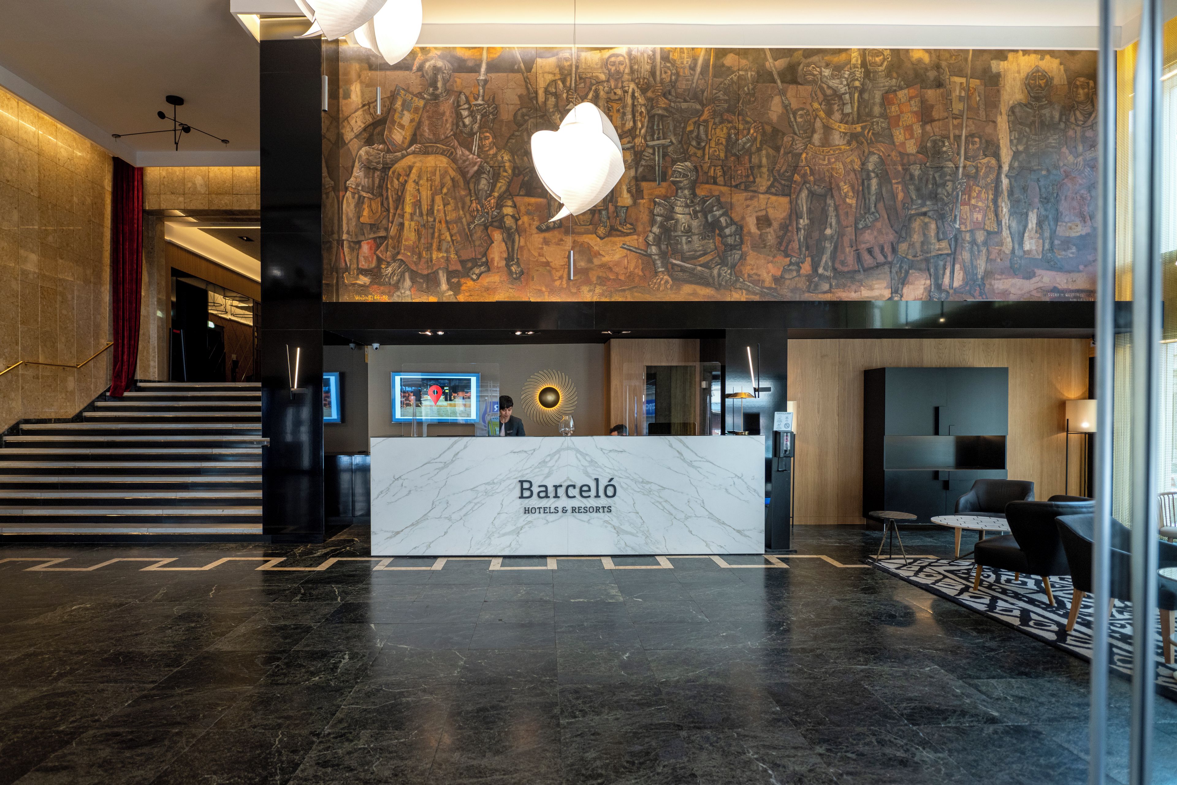 A modern hotel lobby with a marble reception desk, a large mural, and a staircase.
