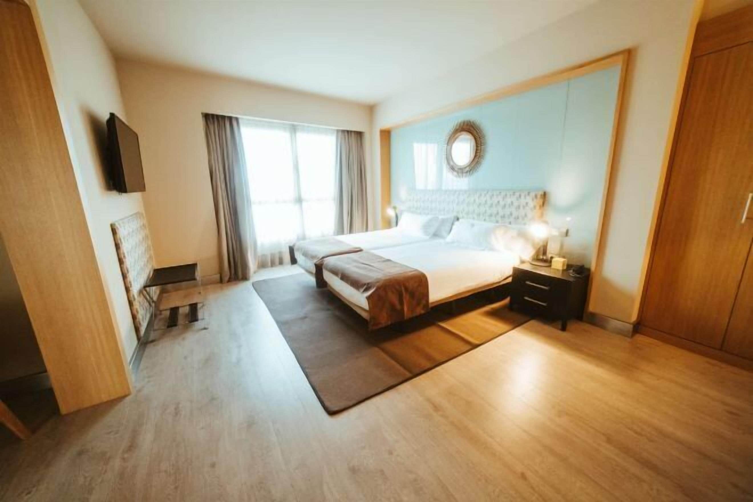 Habitació d'hotel moderna amb un llit gran, una taula de nit, un armari de fusta i un finestral amb cortines.