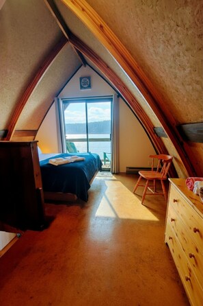 A cozy attic bedroom with a bed, dresser, and chair, featuring a large window with a view of the outdoors. - Jade Resort (Heriot Bay)